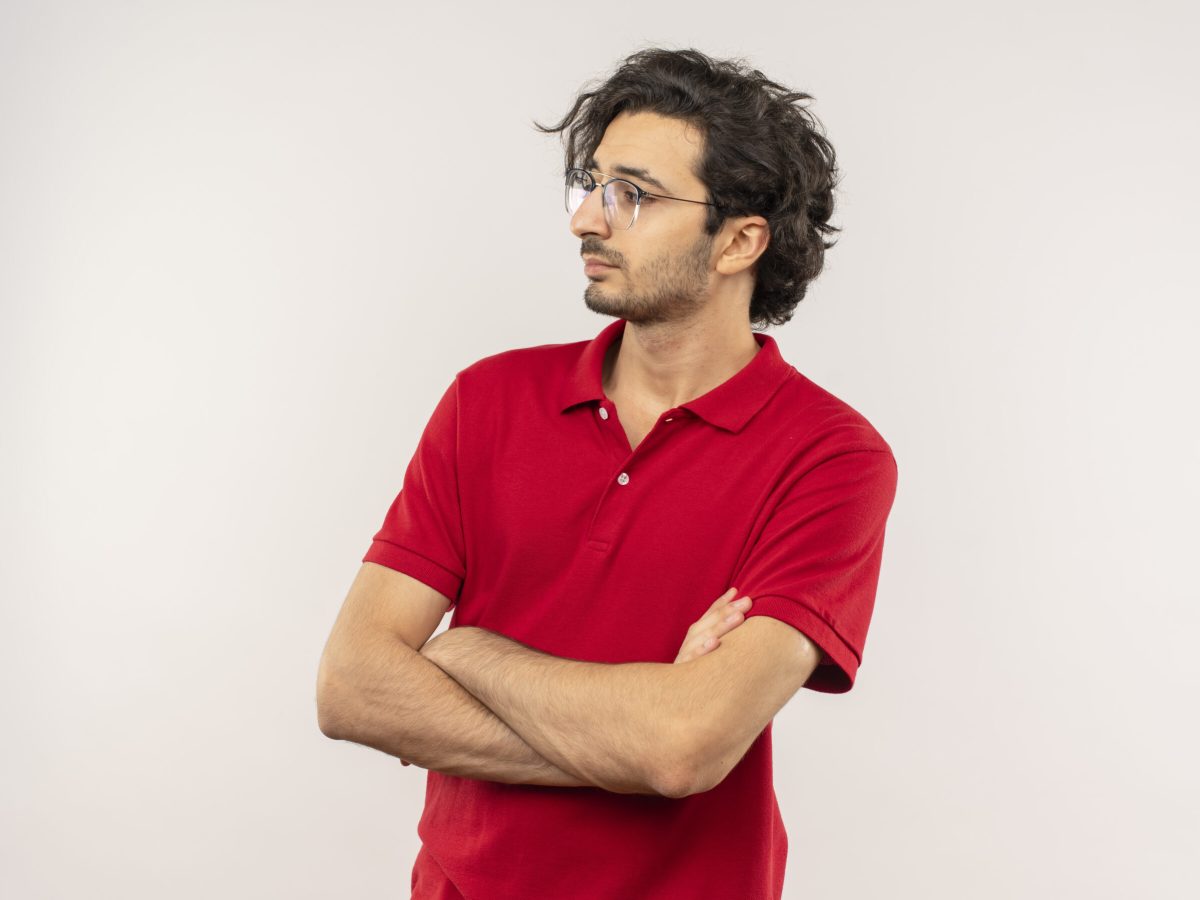 young confident caucasian man in red shirt with optical glasses crosses arms and looks at side isolated on white background with copy space